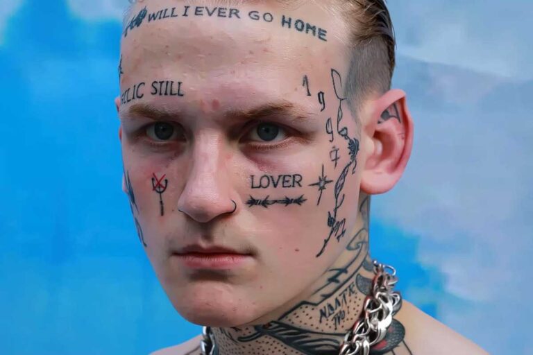 A young man with slick-back dirty-blond hair and icy blue-gray eyes, face covered in fine-line black tattoos