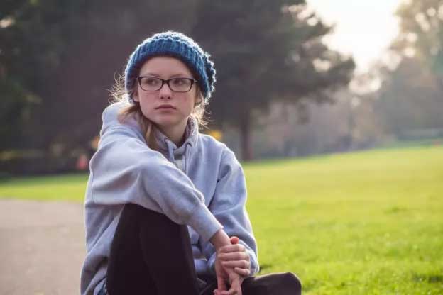 Teenage girl sitting in a park, wearing hooded sweater and beanie, looking as though she is lacking in self-confidence.