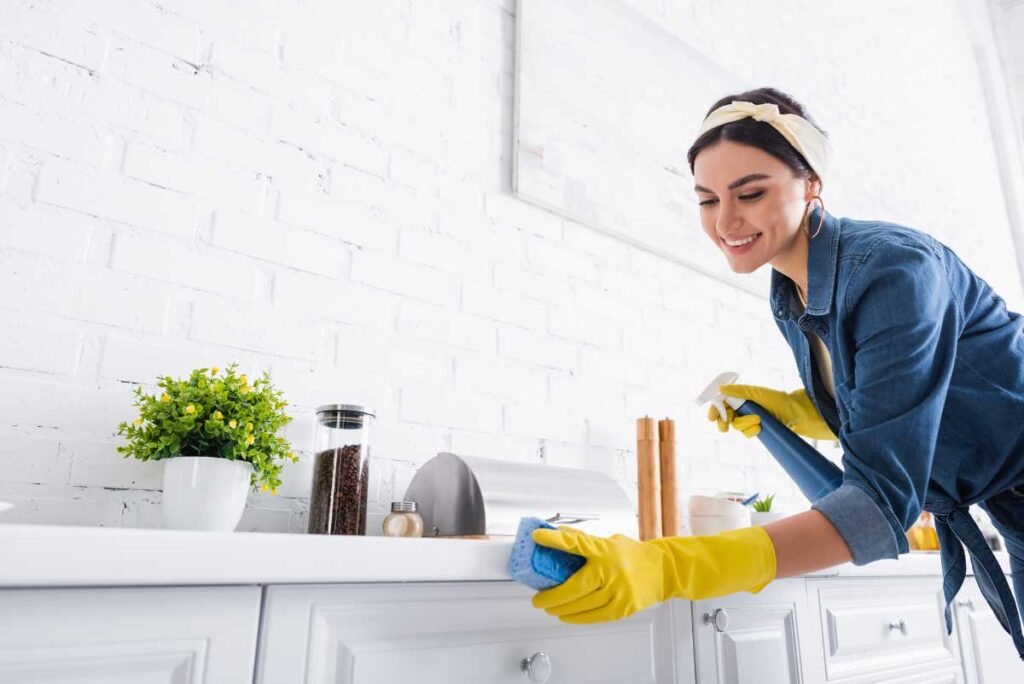 Smiling woman in yellow gloves cleaning kitchen counter, promoting housekeeping services.