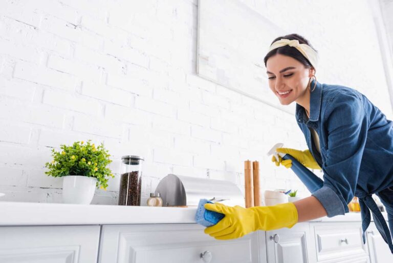 Smiling woman in yellow gloves cleaning kitchen counter, promoting housekeeping services.