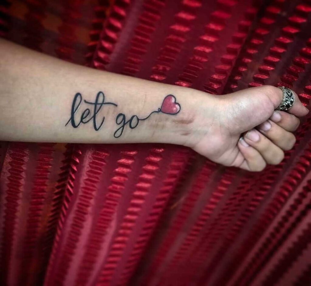 Close-up of a 'let go' tattoo with heart on forearm against red fabric background.