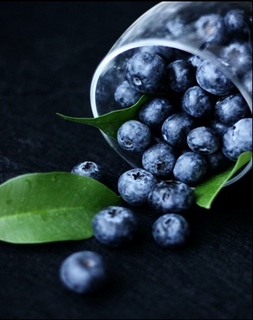 Spilled blueberries from a glass cup with green leaves on a dark background