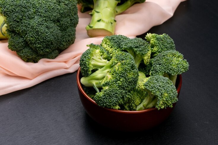 Fresh broccoli florets in a bowl on a dark surface with a peach cloth backdrop.