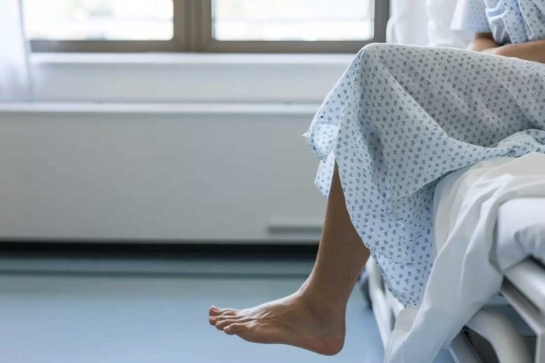 A person in a hospital gown sitting on a bed with legs dangling