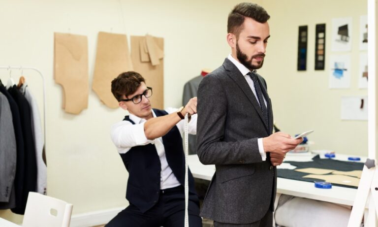 A tailor holding a measuring tape to the back of a man's shoulders. He wears a fitted suit inside a fitting room.