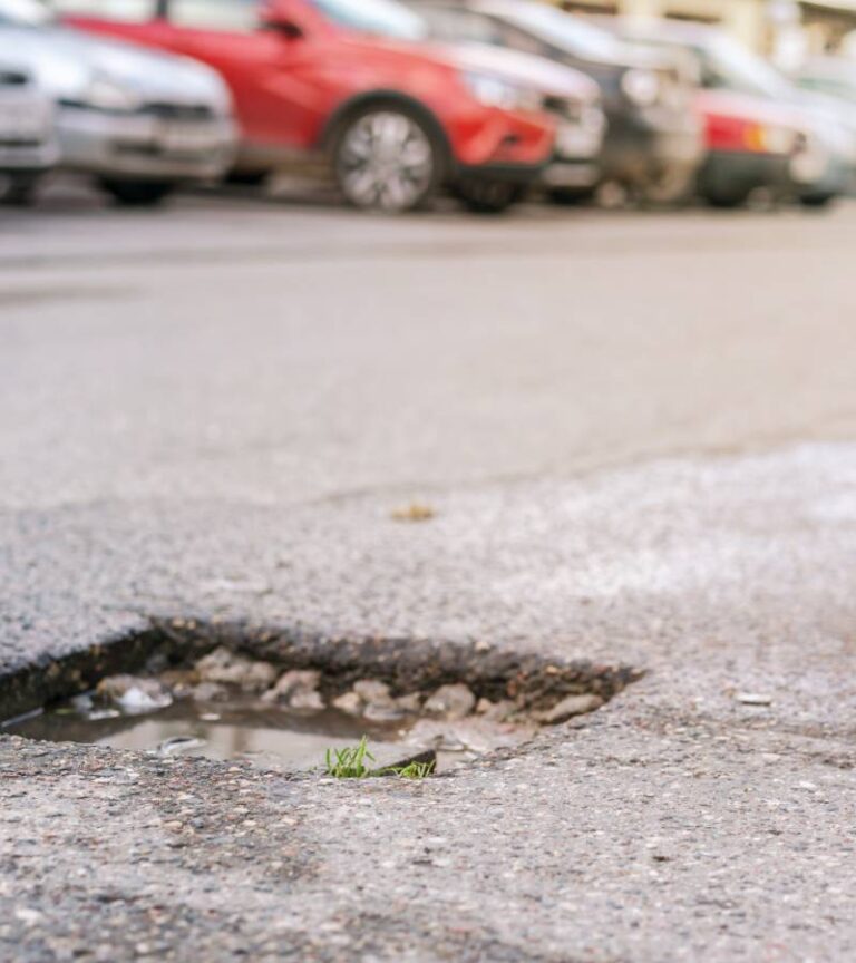A pothole filled with water in a crowded parking lot. There is a line of various cars parked in the parking lot.