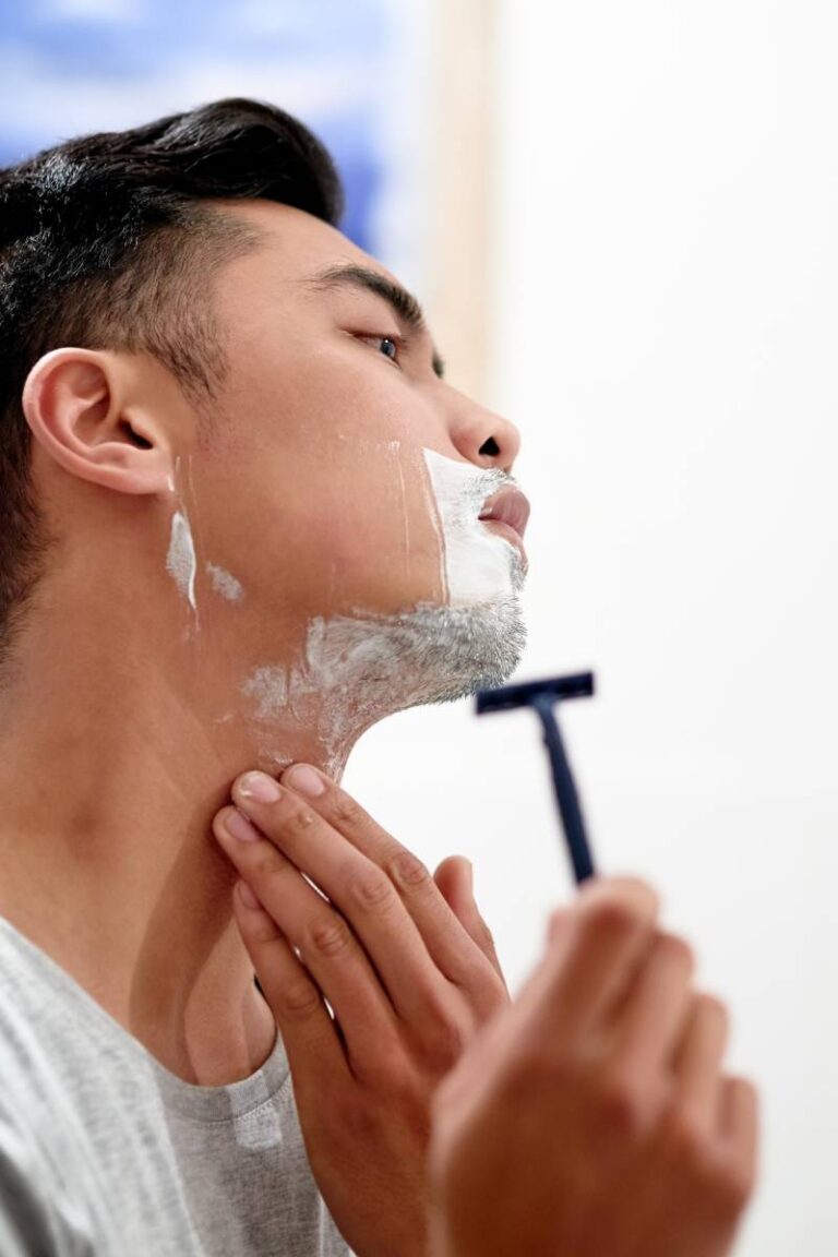 A young man tilting his head to the side so he can more easily shave his jawline and neck with a razor.
