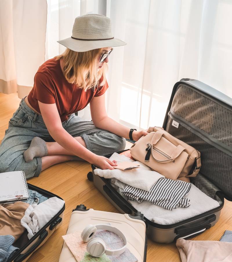 A young woman sitting on the floor surrounded by suitcases. She is packing clothing and accessories into an open suitcase.