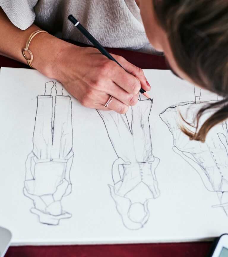 A blonde woman leaning over a large sketchbook on a desk as she draws three fashion design concepts.