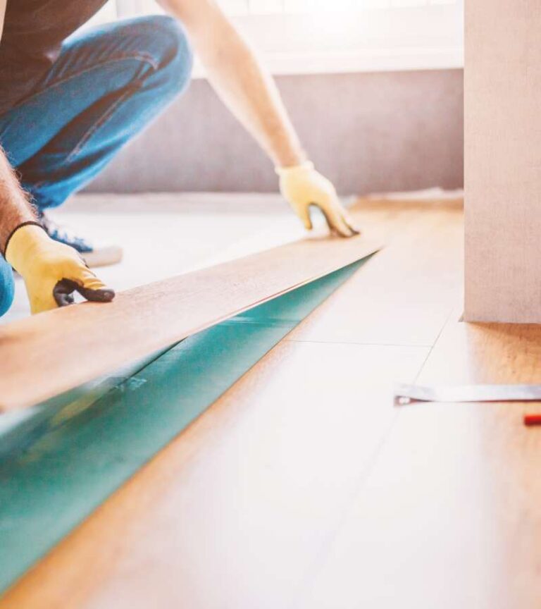 A worker wearing jeans, a black shirt, and yellow gloves lays down new flooring during a home renovation.