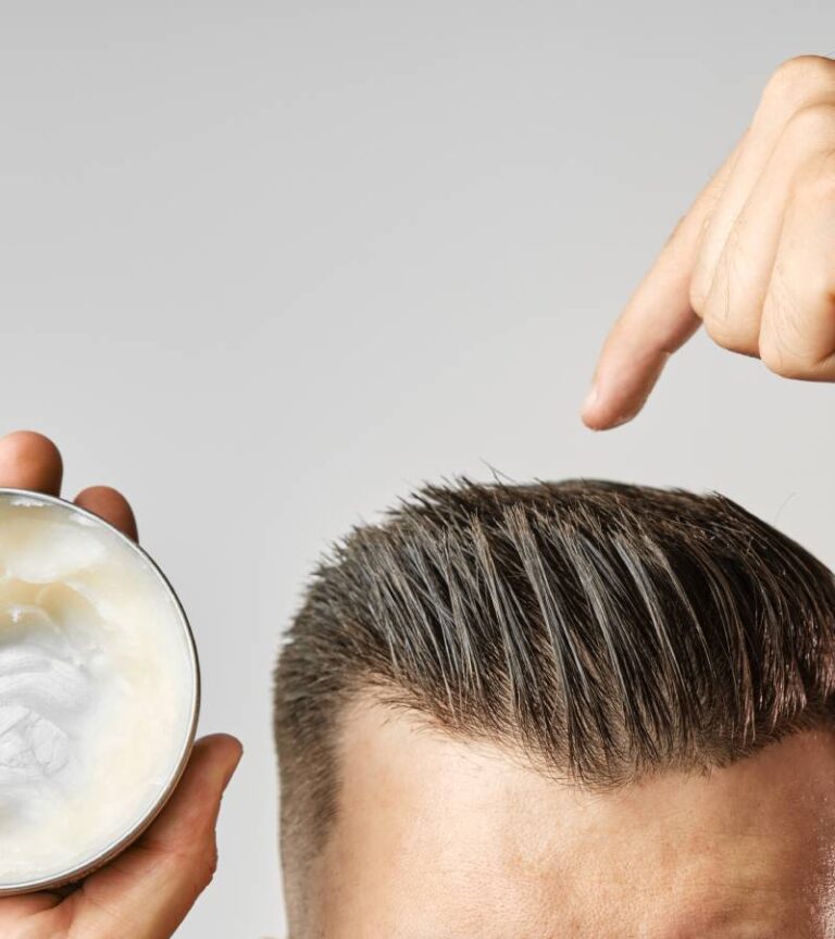 A man is holding an open tub of hair pomade. With his other hand, he's pointing to his recently styled and matte-finished hair.