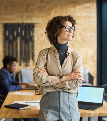 A female business leader stands at the head of the table with her arms crossed and looking out the window.