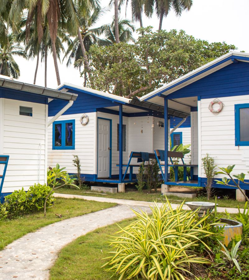 A community of blue and white tiny homes near greenery. Each home has a small porch with a built-in wooden bench.