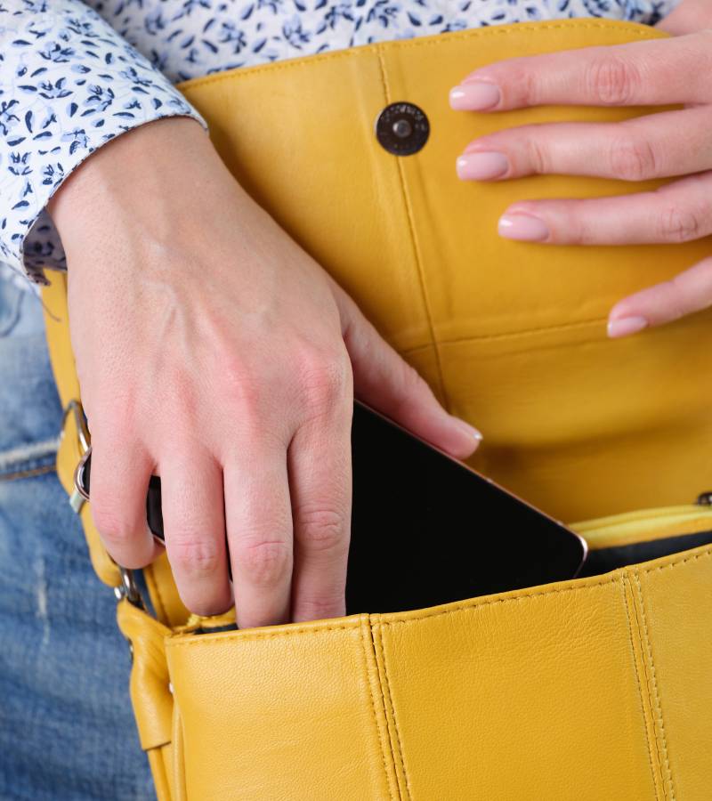 A woman wearing a yellow leather crossbody bag. The bag is open and she is sliding her smartphone inside.