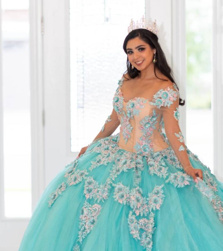 A teenager smiles as she stands by a window and wears teal quinceañera dress with floral applique and sheer sleeves.