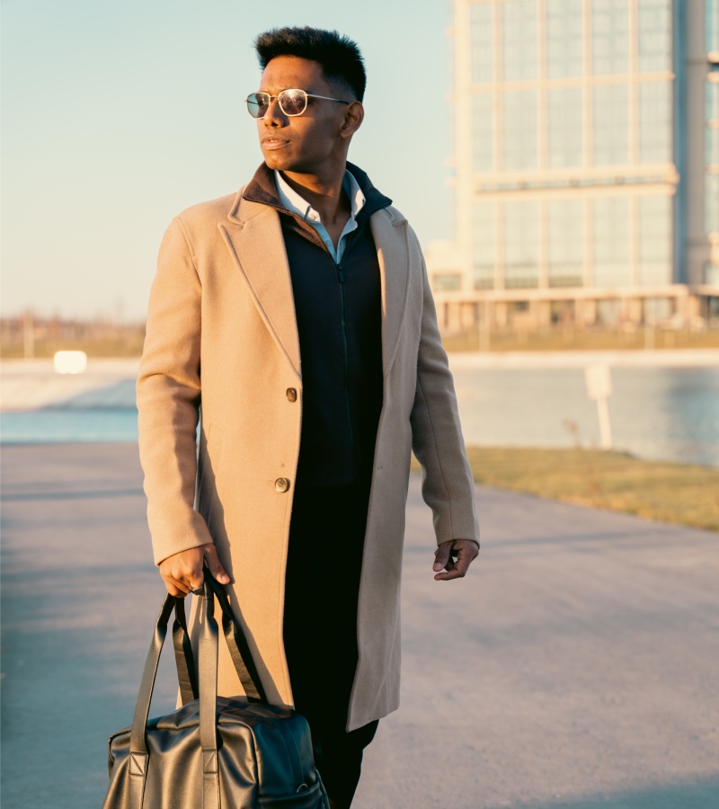 A man is dressed in business professional attire. He is carrying a black bag and wearing a trench coat and sunglasses.