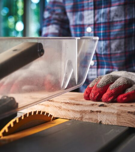 A close-up view of a gloved hand pushing a piece of wood into the blade of a yellow circular table saw.