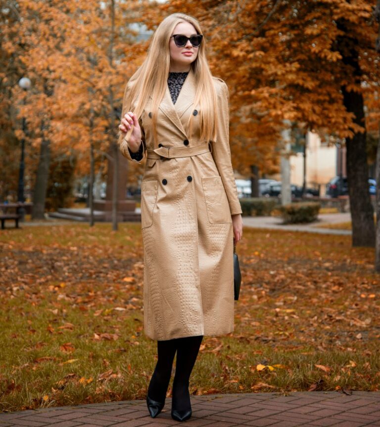 A blonde woman stands among fall leaves wearing black sunglasses, a beige trenchcoat, and black boots.
