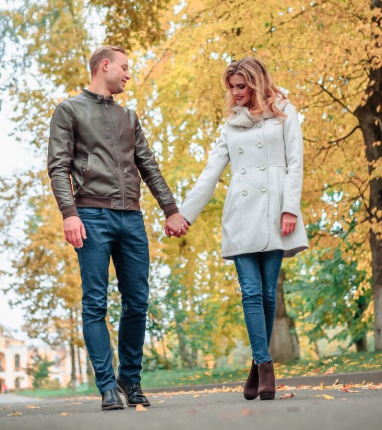 A young man and woman hold hands as they take a walk in a park on an autumn day to look at the changing leaves.