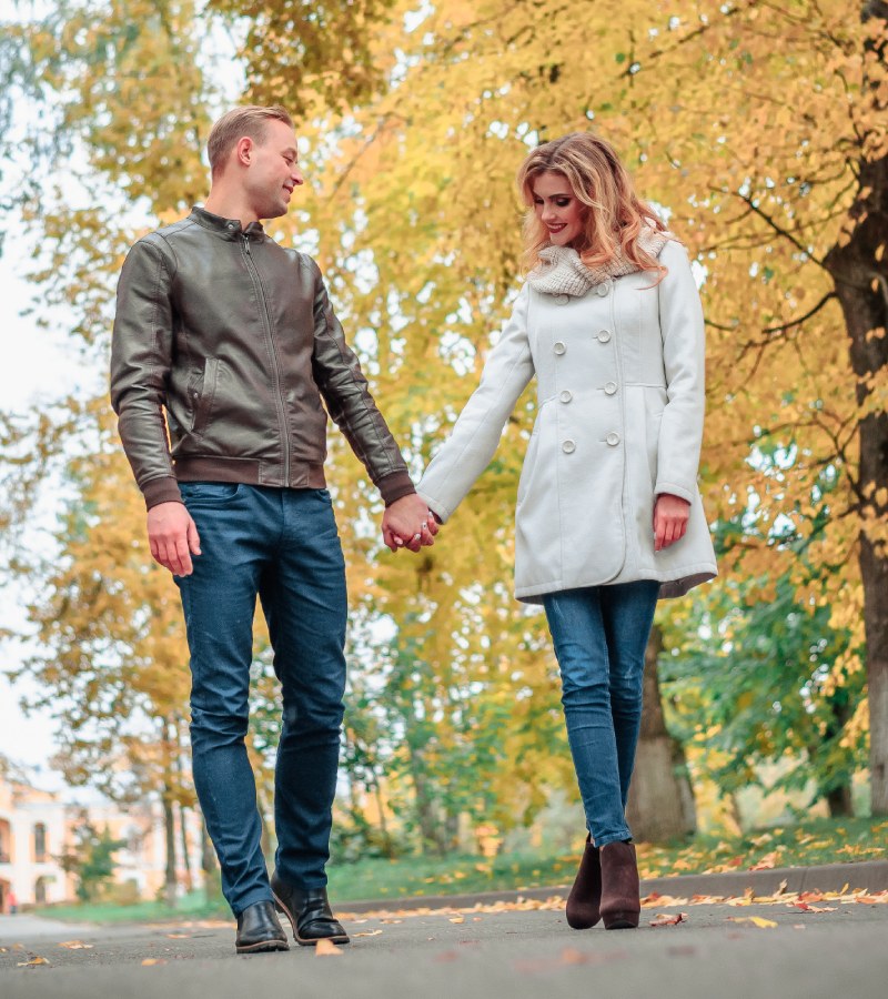 A young man and woman hold hands as they take a walk in a park on an autumn day to look at the changing leaves.