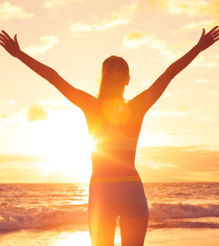 A happy woman rejoices with her arms waving in the air while the sun sets. She's standing on the beach.