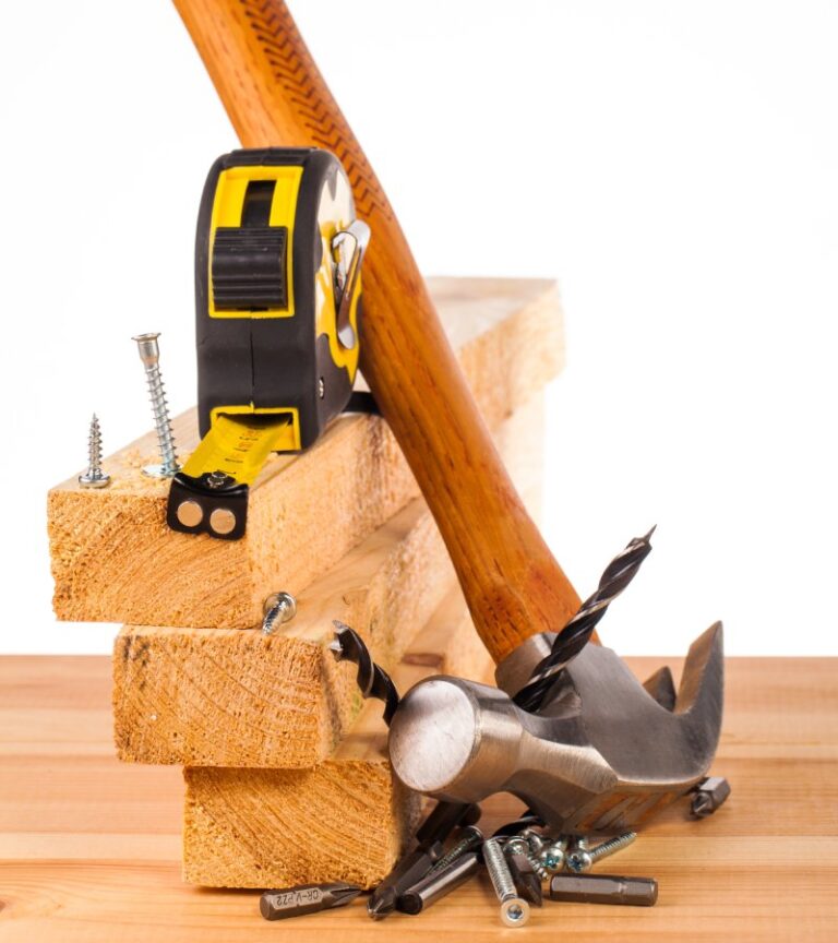 Three pieces of wood sit on top of each other with a measuring tape on top and a hammer and nails resting on the side.