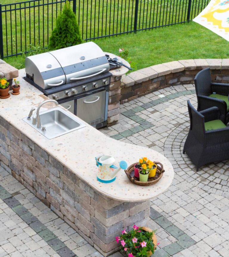Overhead view of an outdoor kitchen next to table and chairs. Potted plants and a water can are on the kitchen island.