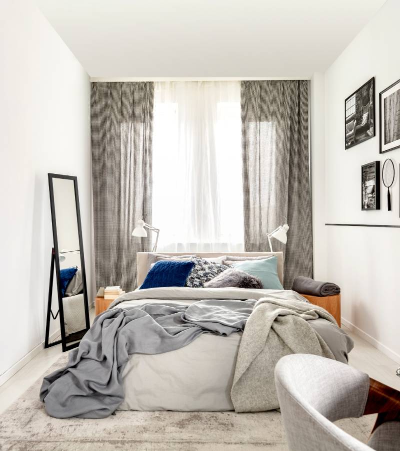 A small white bedroom contains a bed, a tall, black mirror, and a small table with a chair. One wall includes photos.