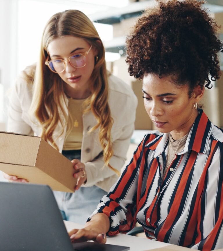 Two young women look at an open laptop. One is sitting in a black chair, and the other stands while holding a cardboard box.