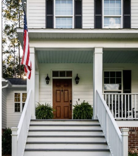 A home has white siding, black shutters, a wooden front door, and a large front porch. An American flag sways in the wind.