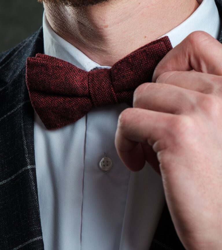 A close-up of a person touching their bow tie. The bow tie is red, and the suit jacket is dark with patterns.