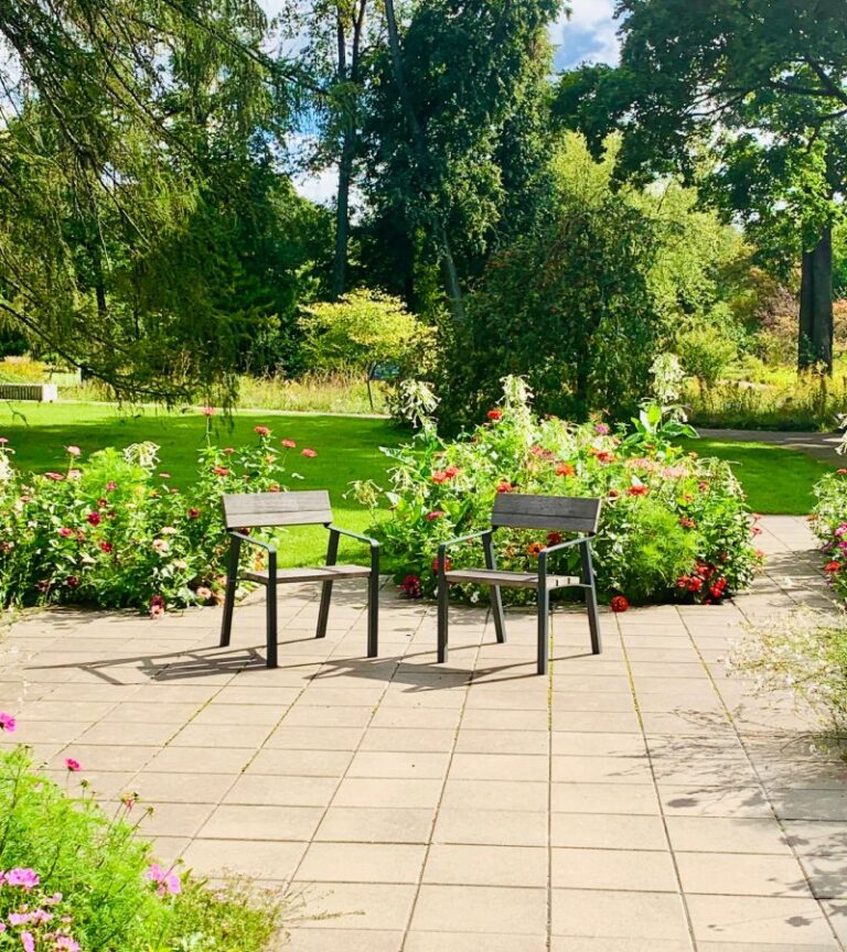 Two metal chairs with wooden seats sit on a concrete patio. Lush flowers and green grass surround the patio.