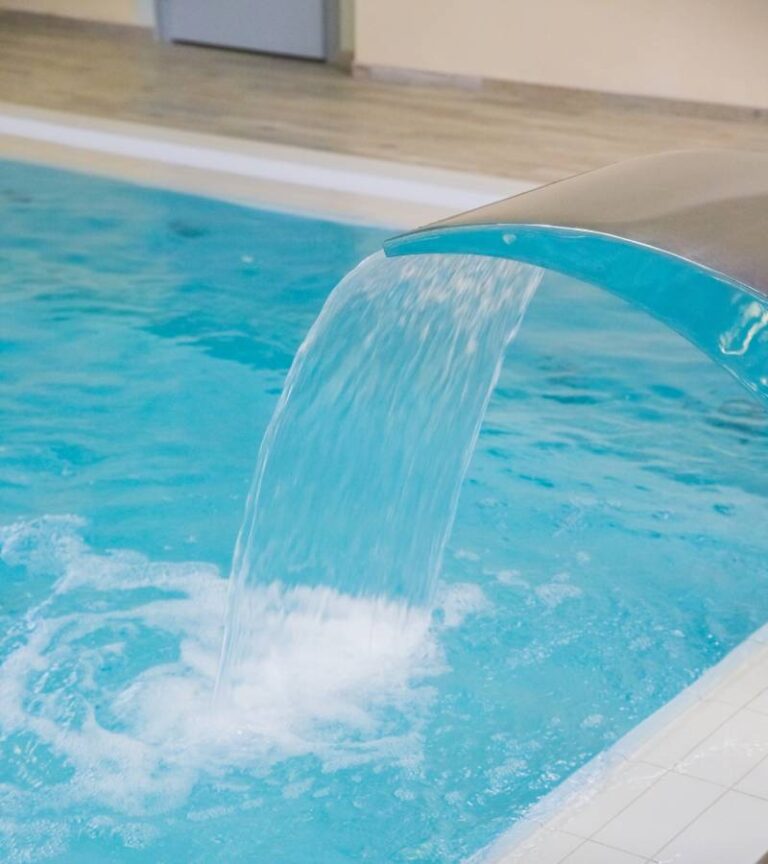 A small waterfall flows into a pool filled with crystal clear, blue water. The pool is surrounded by white and tan tile.