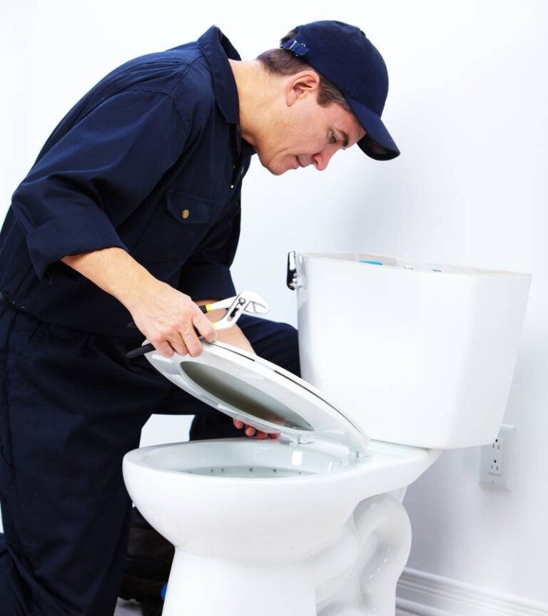 A man in a navy blue jumpsuit and hat kneels next to a white toilet with his colorful tools on the floor.