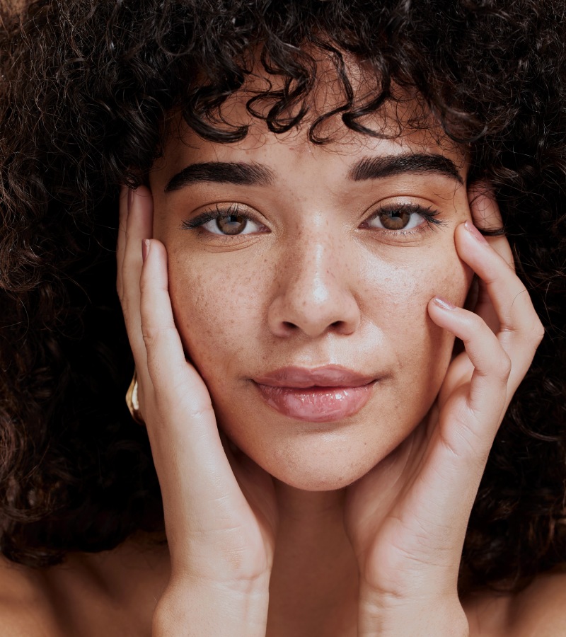 A young woman with dark curly hair and dark brown eyes has her hands on her face. Her face is covered in freckles.