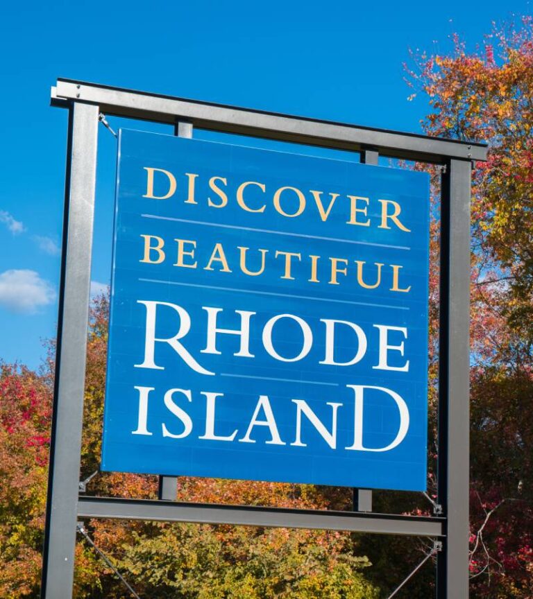 A sign displaying "Discover Beautiful Rhode Island" stands against a backdrop of a blue sky and trees with fall foliage.