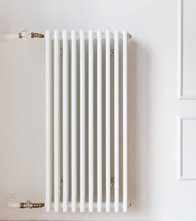 A white radiator sits on a white wall with two white pipes attached. One is attached at the top and one on the bottom.