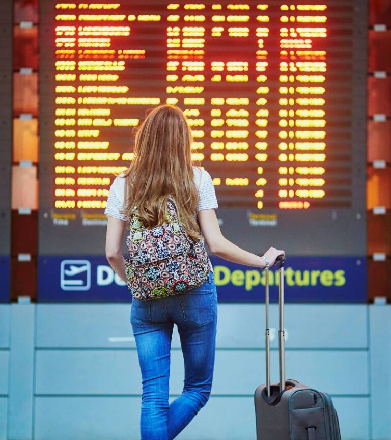 A woman stands in front of a flight information board. She wears a backpack and rests her hand on a suitcase.