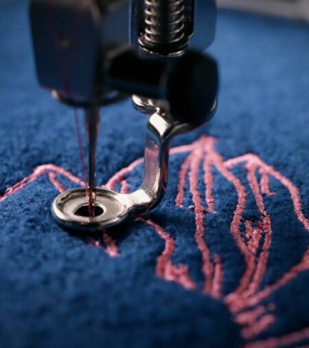 A close-up of an embroidery machine stitching a pink floral design on a navy blue wool-like textured fabric.