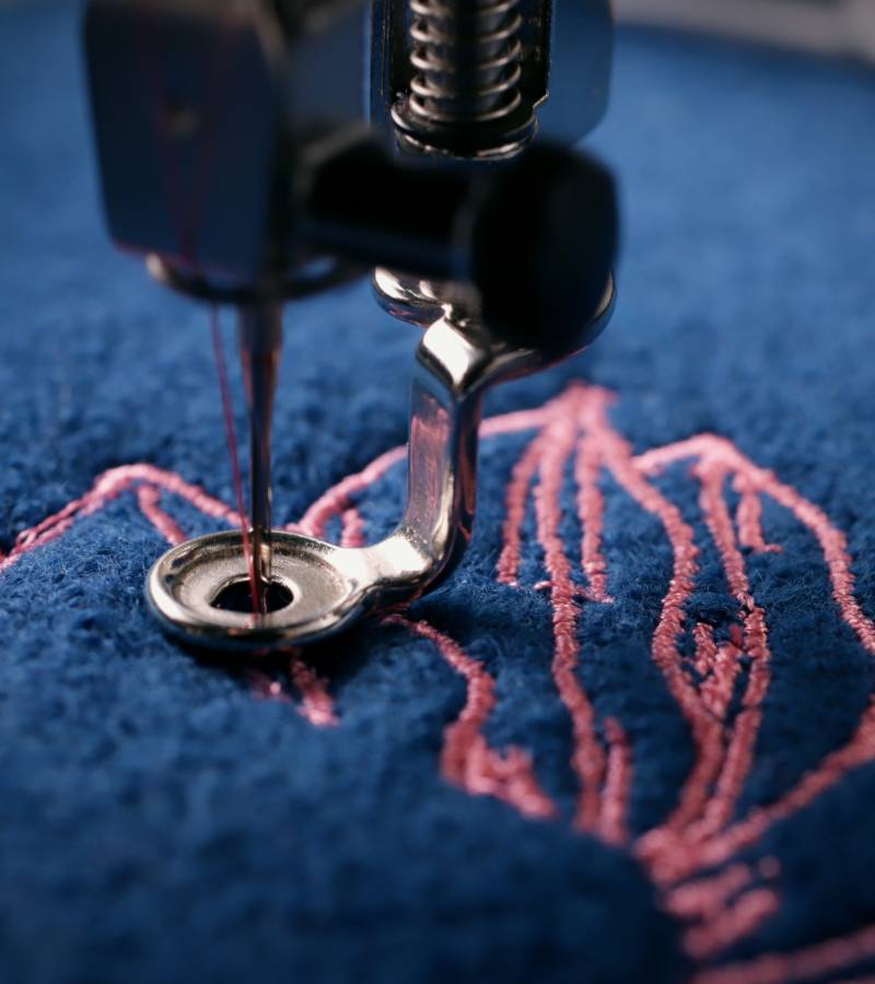 A close-up of an embroidery machine stitching a pink floral design on a navy blue wool-like textured fabric.