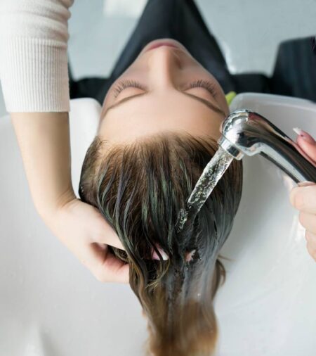 A woman lays back, allowing her hair to fall into a white sink. A person uses a silver sprayer to wet her hair.