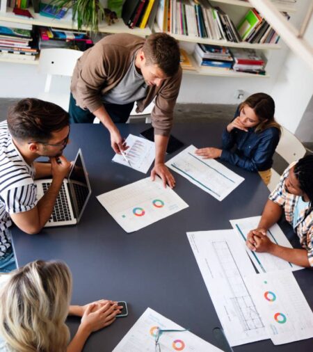 Five people gather around a large, black table that contains various papers with graphs. The people are looking at the papers.