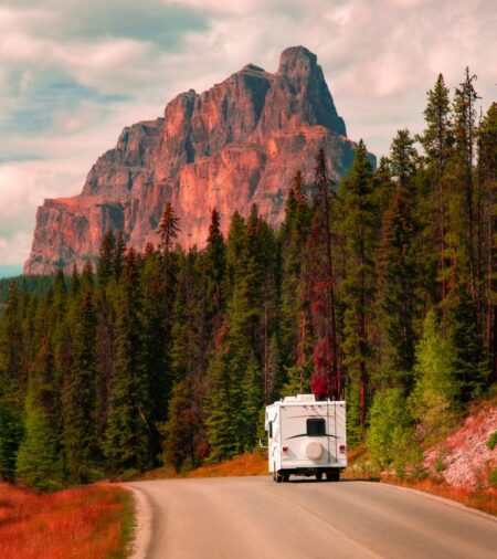 A white RV drives down an empty road while the sunset creates a warm pink hue on the mountains in the background.