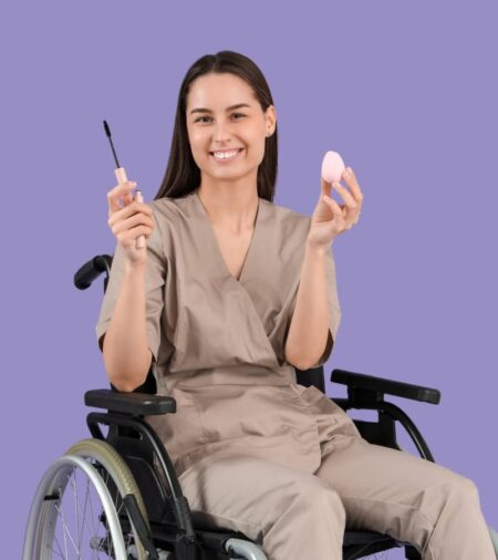 A female wheelchair user holding a mascara wand and makeup sponge. She's smiling and sitting in front of a purple backdrop.
