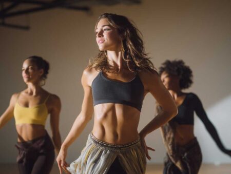 Three dancers in a studio wearing crop, tank, and long sleeve dance tops, mid movement and confident.
