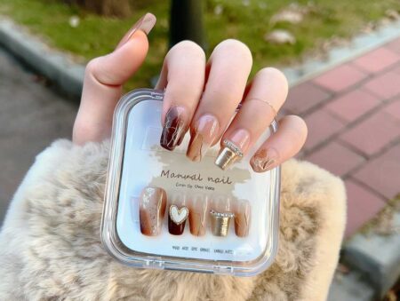 Close-up photo of a left hand, wearing a beige faux-fur coat sleeve, holding a clear square case of press-on nails