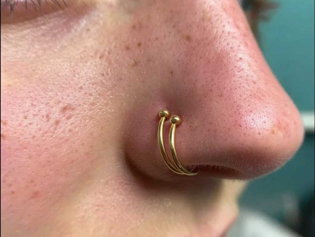 Close-up side view of a nose with two small gold hoops stacked in a single nostril piercing, highlighting realistic skin texture and freckles.