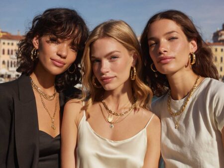 Three models in a blazer, slip dress, and tee showcase wholesale jewelry, chunky gold hoops, pearl drops, enamel bangles, and layered zodiac necklaces
