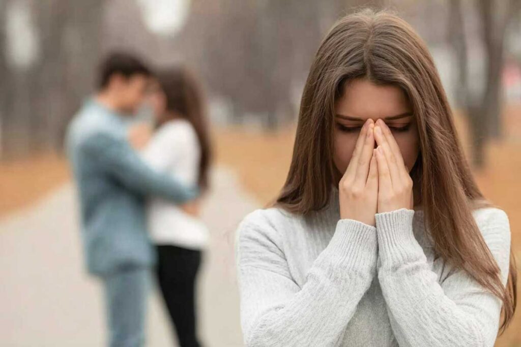 Heartbroken young woman in the foreground, long straight brown hair, light gray sweater, covering her face with both hands in despair