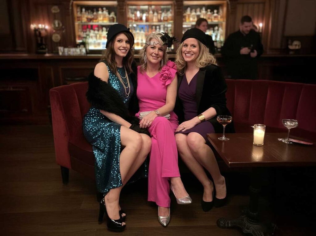 Three women dressed in elegant speakeasy-style outfits sitting on a red velvet sofa in a dimly lit bar, smiling with cocktails on the table.